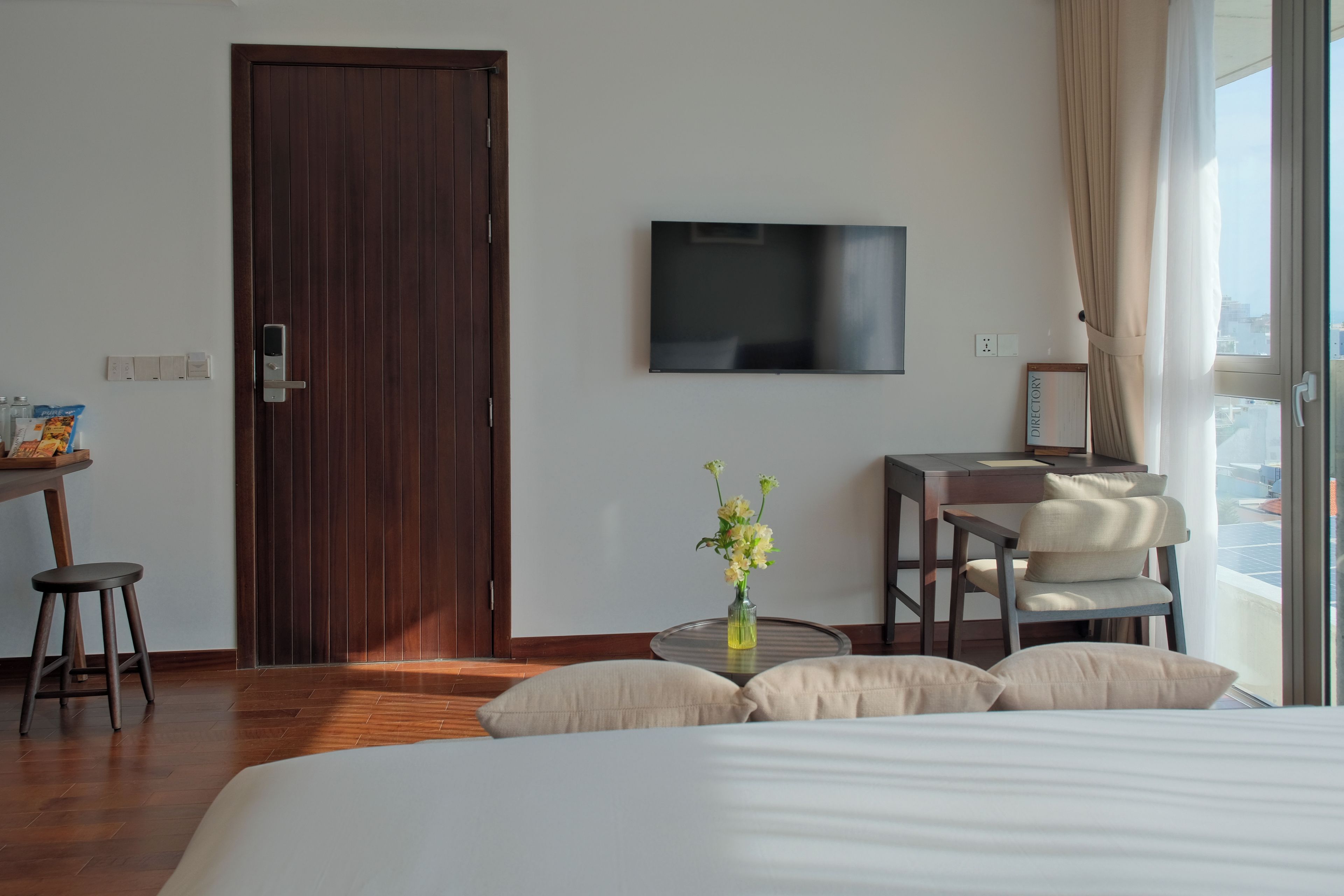 A hotel room with a wooden door, a flat-screen TV mounted on the wall, a desk with a chair, a bed with pillows, and a window with curtains.