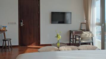 A hotel room with a wooden door, a flat-screen TV mounted on the wall, a desk with a chair, a bed with pillows, and a window with curtains.