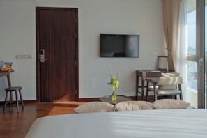 Une chambre d’hôtel avec une porte en bois, un téléviseur à écran plat fixé au mur, un bureau avec une chaise, un lit avec des oreillers et une fenêtre avec des rideaux.