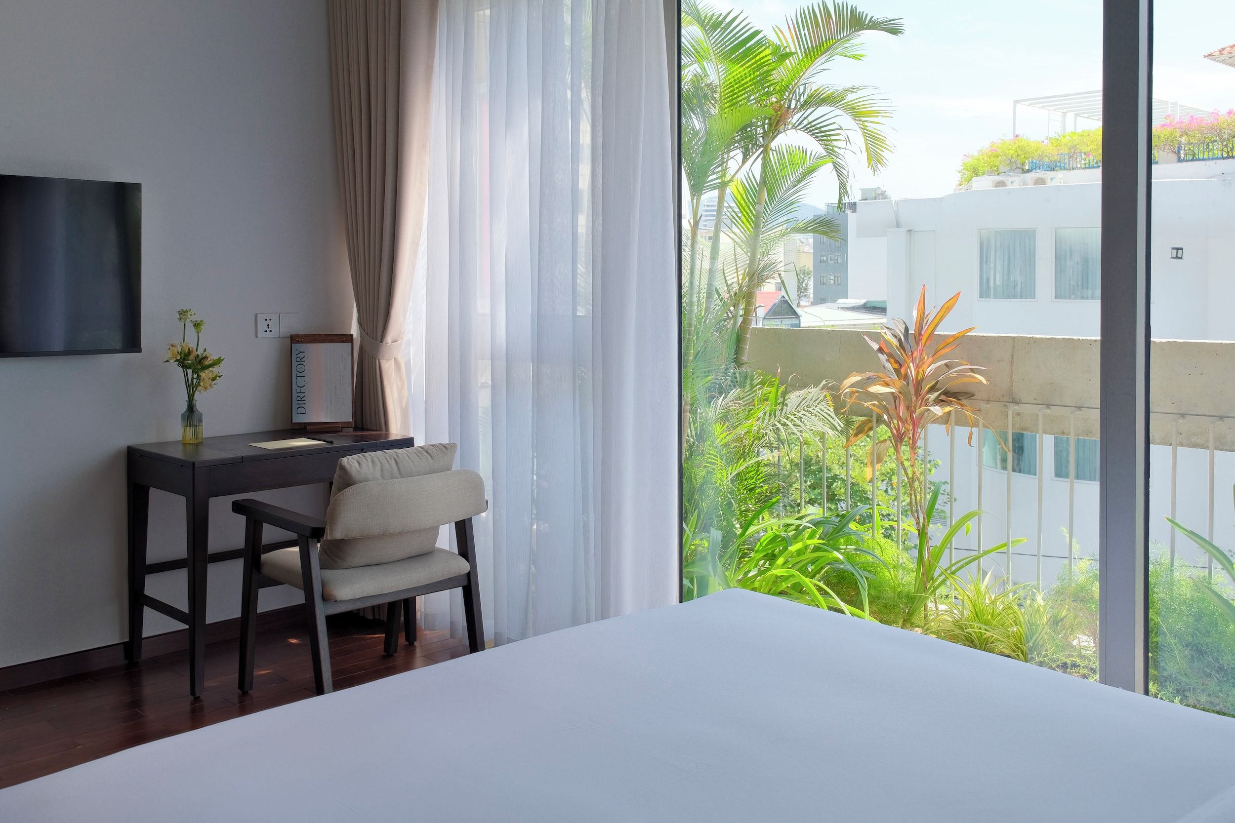 A hotel room with a bed, a desk with a chair, a TV mounted on the wall, and a view of greenery and buildings outside.