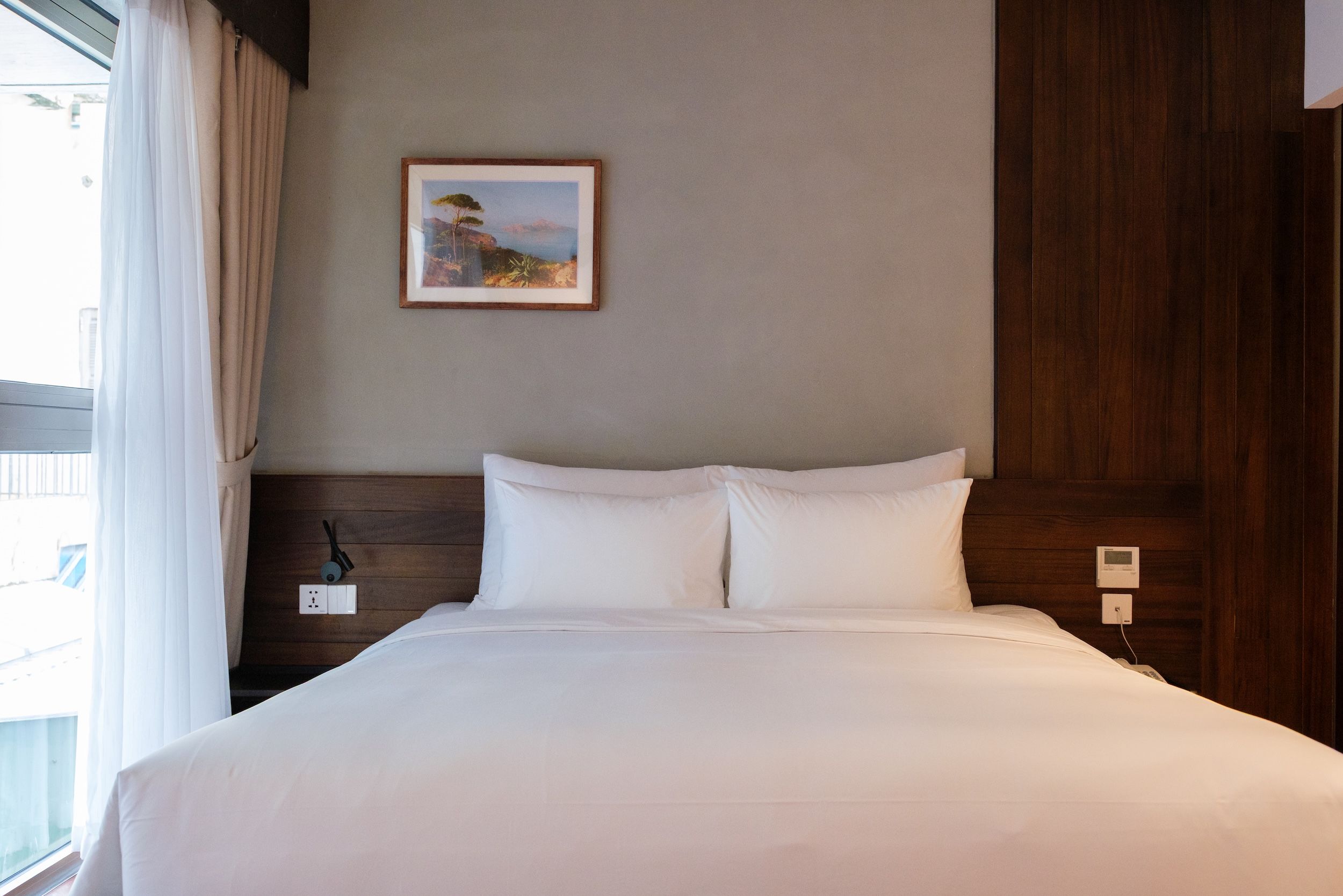 A hotel room with a bed, a nightstand, a power outlet, and a painting on the wall.