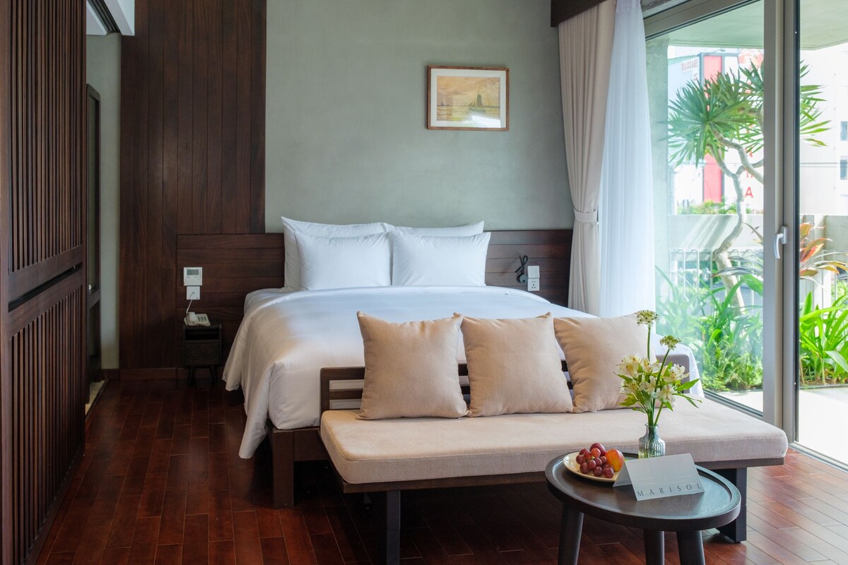 Quarto de hotel com cama, mesas de cabeceira, uma pequena mesa com cartão de nome e vista para o verde lá fora.