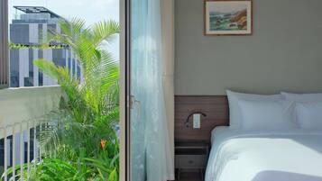 A hotel room with a bed, a nightstand, a wall-mounted lamp, a painting, and a view of a building and greenery outside.