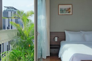 Une chambre d’hôtel avec un lit, une table de chevet, une lampe fixée au mur, un tableau et une vue sur un bâtiment et la verdure environnante.