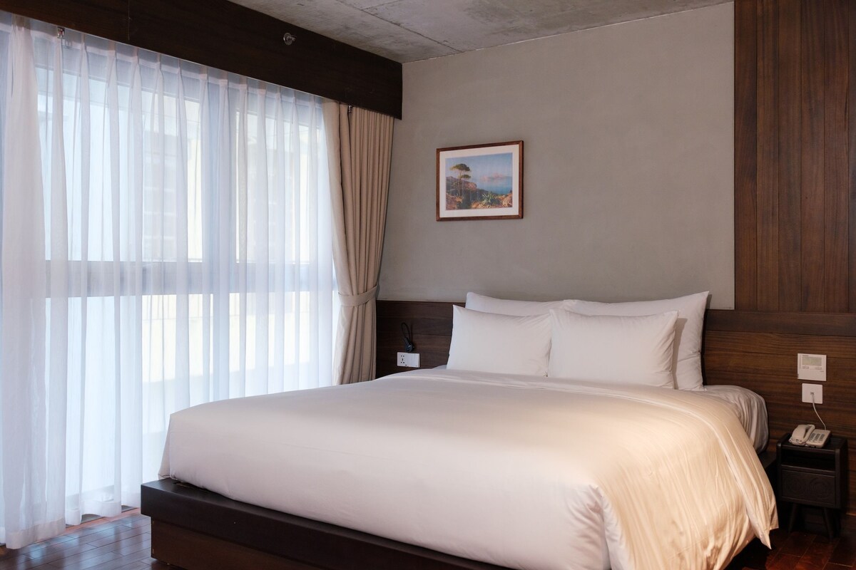 Quarto de hotel com uma cama grande, mesa de cabeceira, um quadro na parede e cortinas leves.