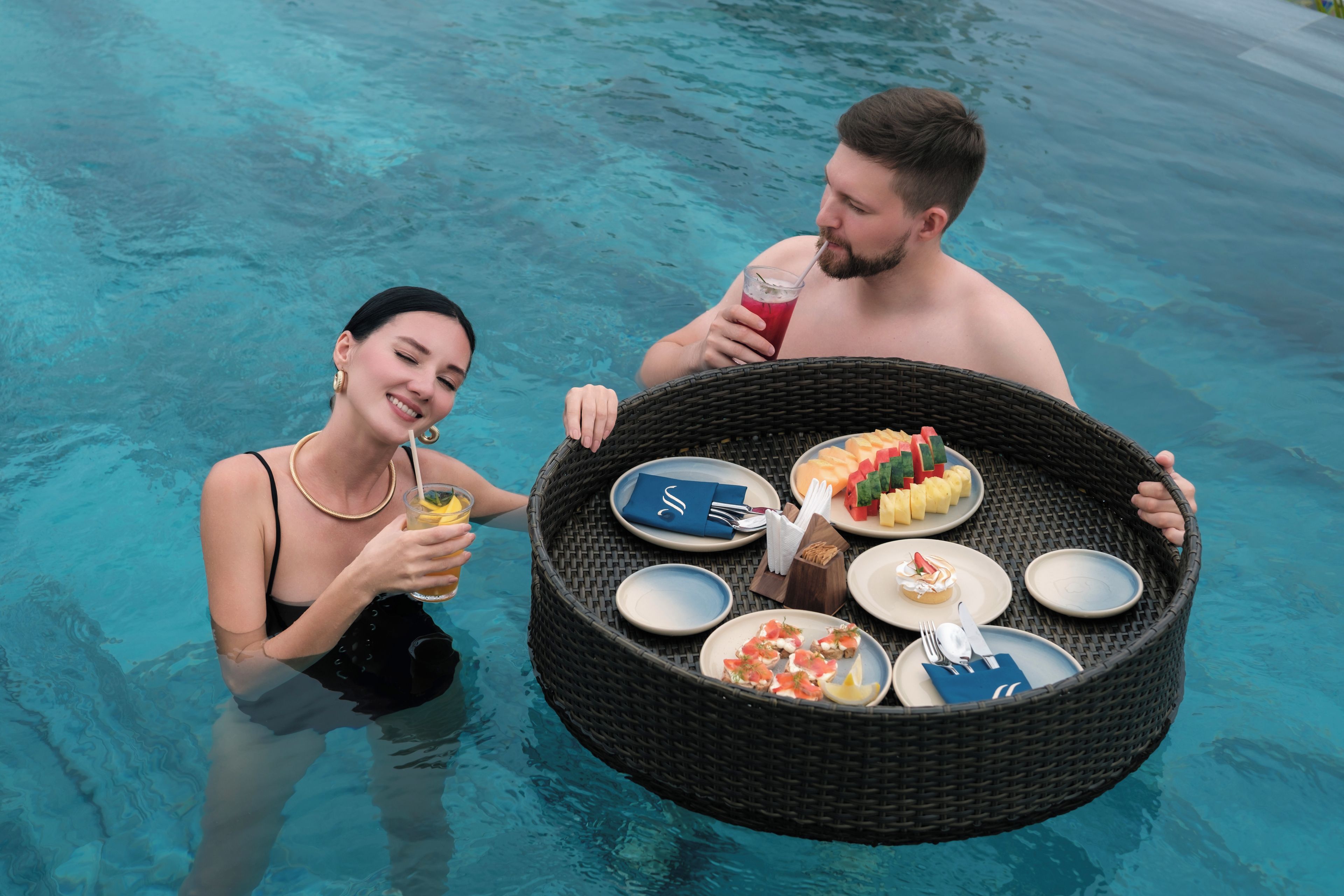 Two people in a swimming pool holding a large wicker basket with plates of food and drinks.