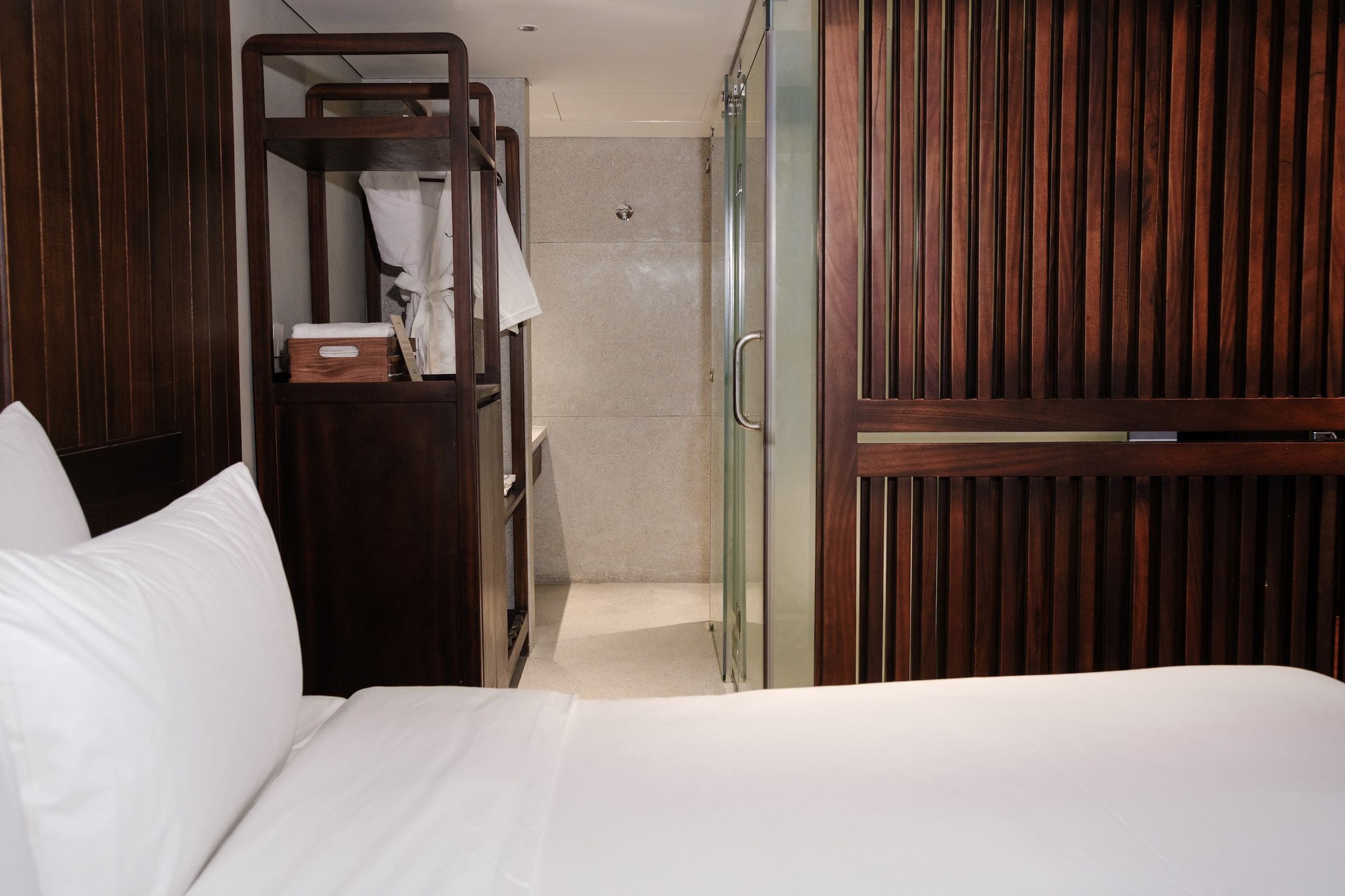 A hotel room with a bed, a wooden wardrobe, a glass shower enclosure, and a white towel.