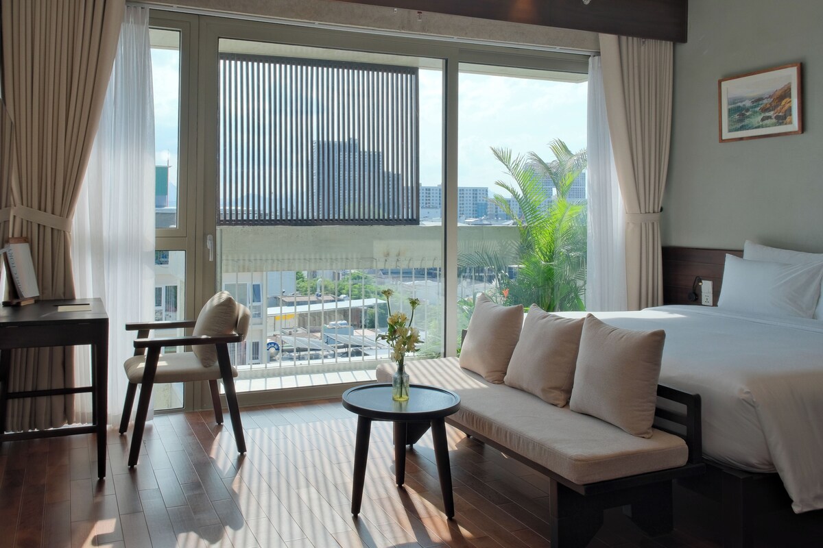 Quarto de hotel com cama, sofá, escrivaninha e vista da paisagem urbana através de grandes janelas.