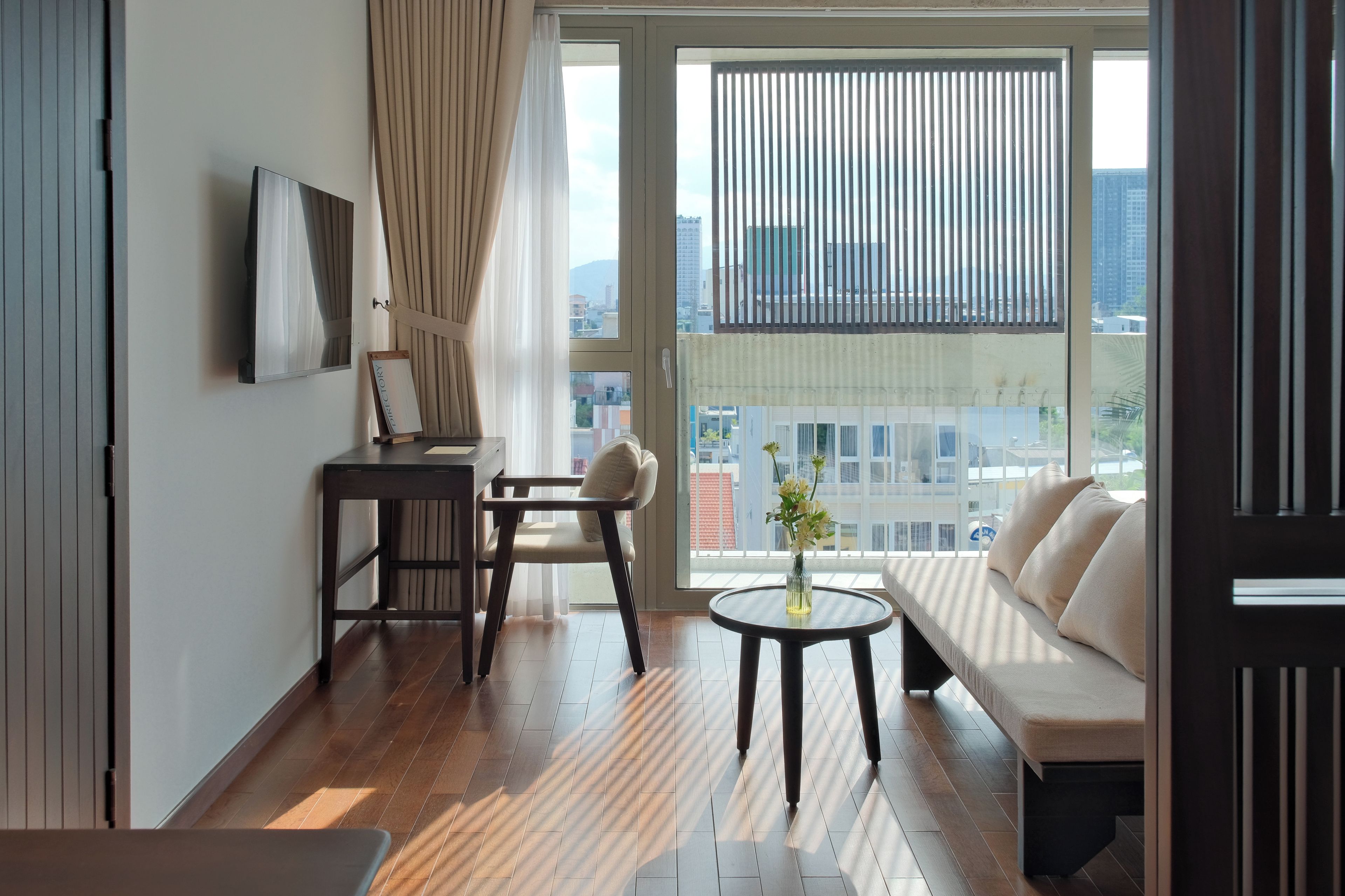 Une chambre d’hôtel moderne avec un plancher en bois, un canapé, une petite table surmontée d’un vase, une chaise, un bureau avec une télévision et une vue sur la ville grâce à de grandes fenêtres.