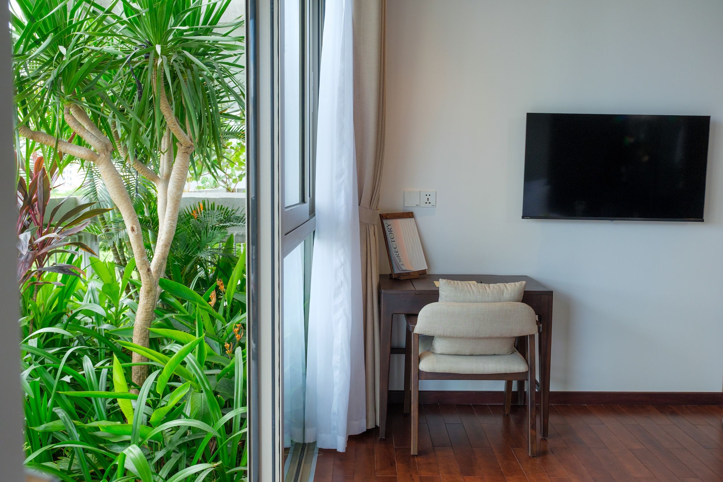 A room with a large window, a desk with a chair, a flat-screen TV mounted on the wall, and a potted plant.