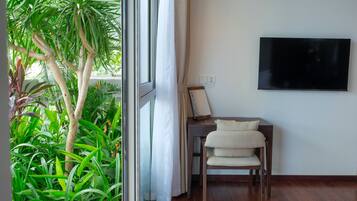 A room with a large window, a desk with a chair, a flat-screen TV mounted on the wall, and a potted plant.