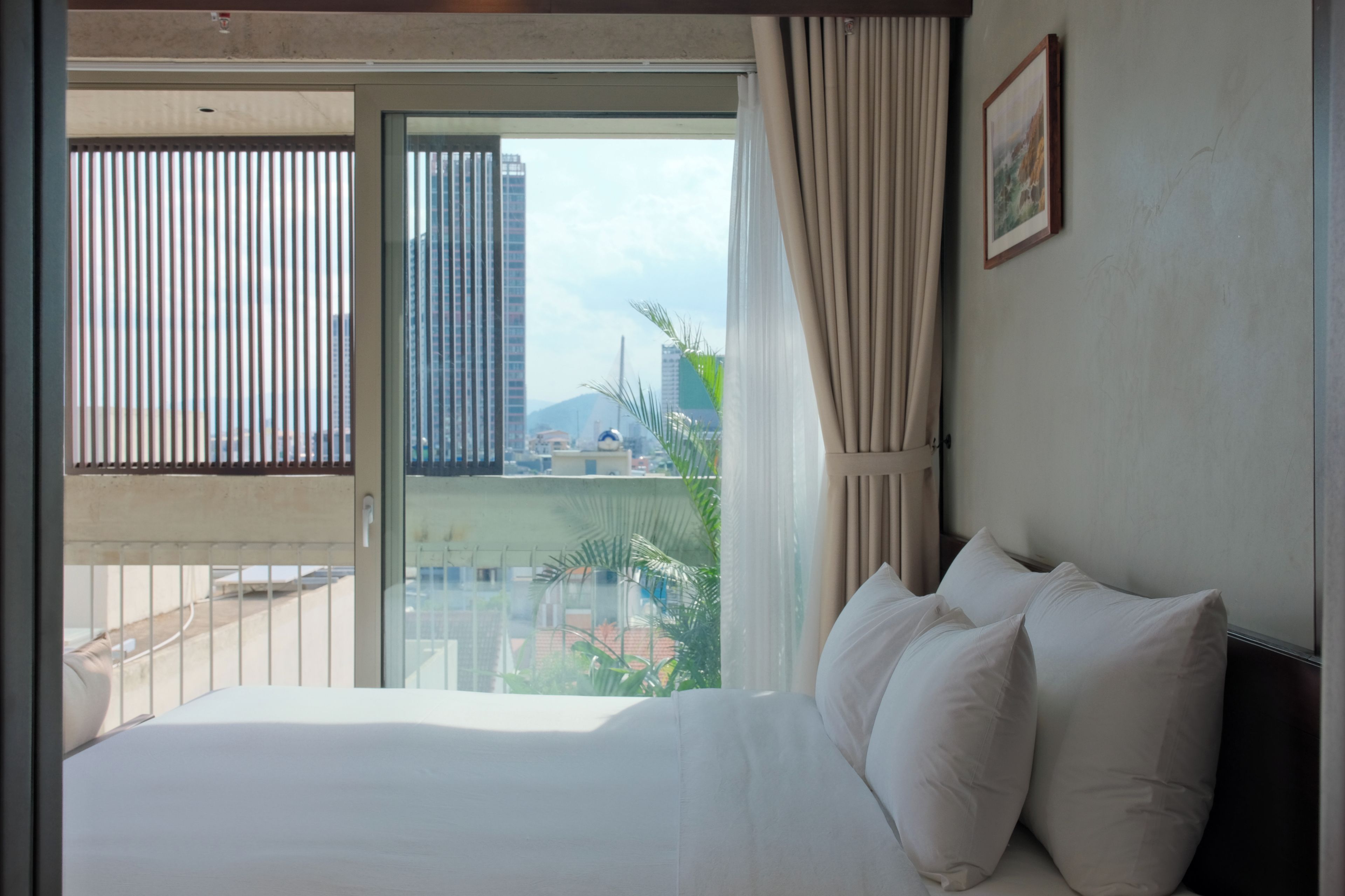 A hotel room with a bed, a window with curtains, and a view of buildings and greenery outside.