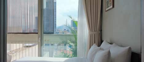 A hotel room with a bed, a window with curtains, and a view of buildings and greenery outside.