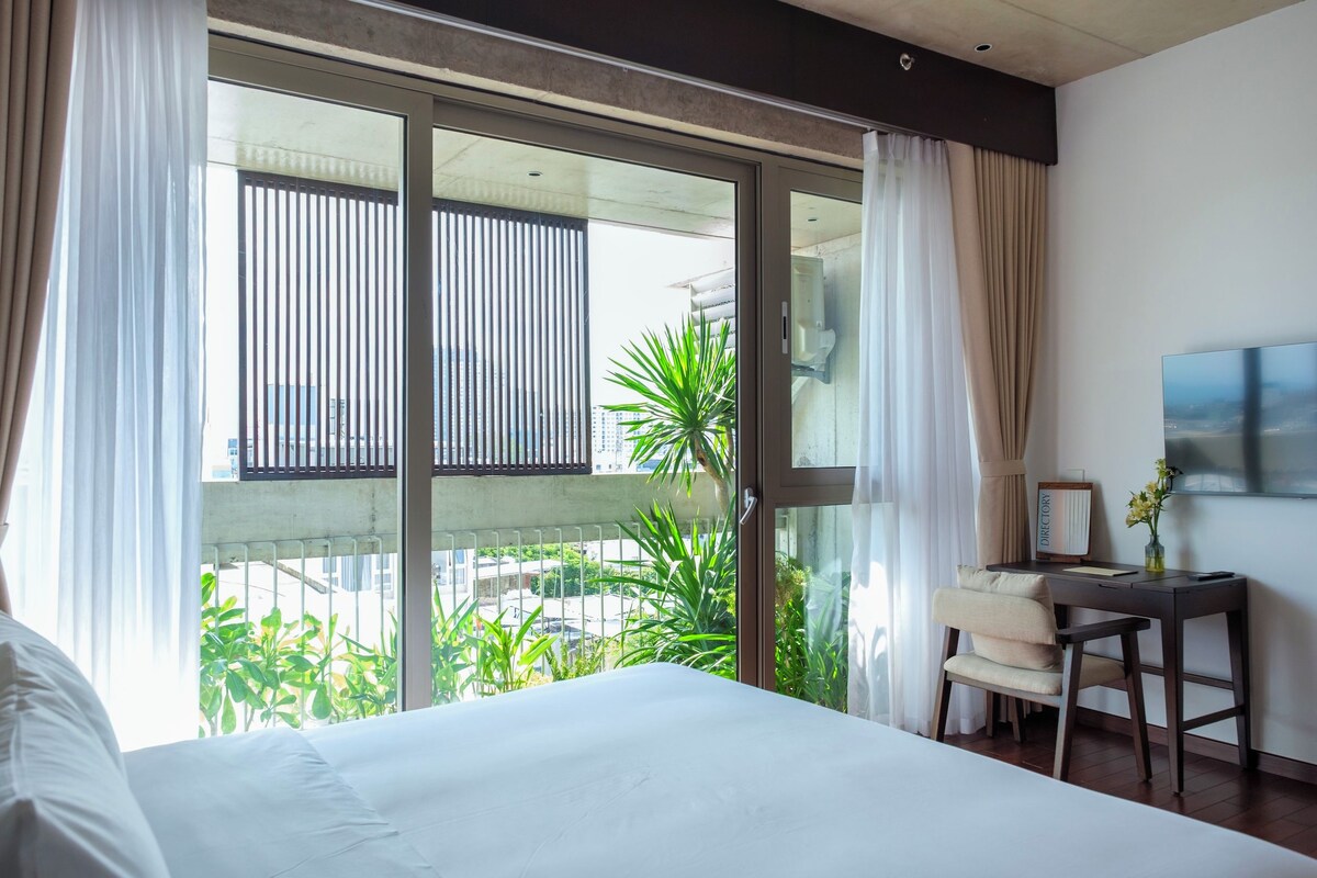 Quarto de hotel com uma cama grande, uma escrivaninha com cadeira e vista para áreas verdes e edifícios pela janela.