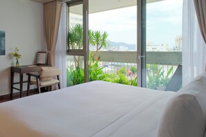 Quarto de hotel com cama, escrivaninha, cadeira, um vaso com flores e vista da cidade através de grandes janelas.
