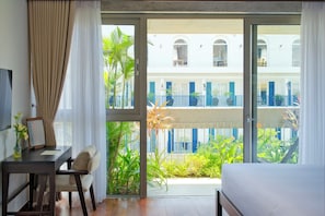 A hotel room with a bed, a desk, a chair, and a view of a building with balconies. - Marisol Hotel (Da Nang)