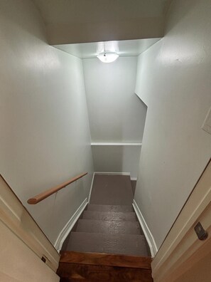 Un escalier étroit doté d’une rampe en bois, menant à un palier sur lequel est fixée une lampe.