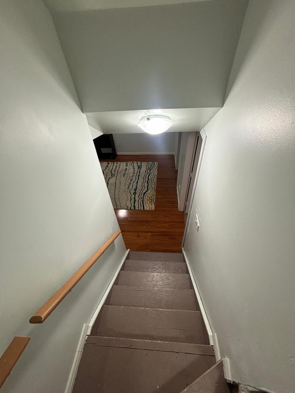 Una escalera estrecha con escalones de madera y paredes blancas, que conduce a una habitación con un tapete estampado y una lámpara en el techo.