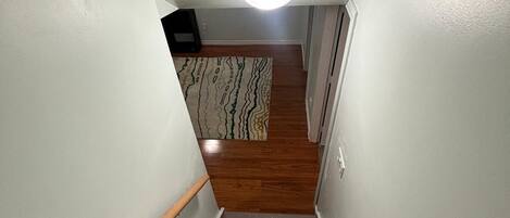 Una escalera estrecha con escalones de madera y paredes blancas, que conduce a una habitación con un tapete estampado y una lámpara en el techo.