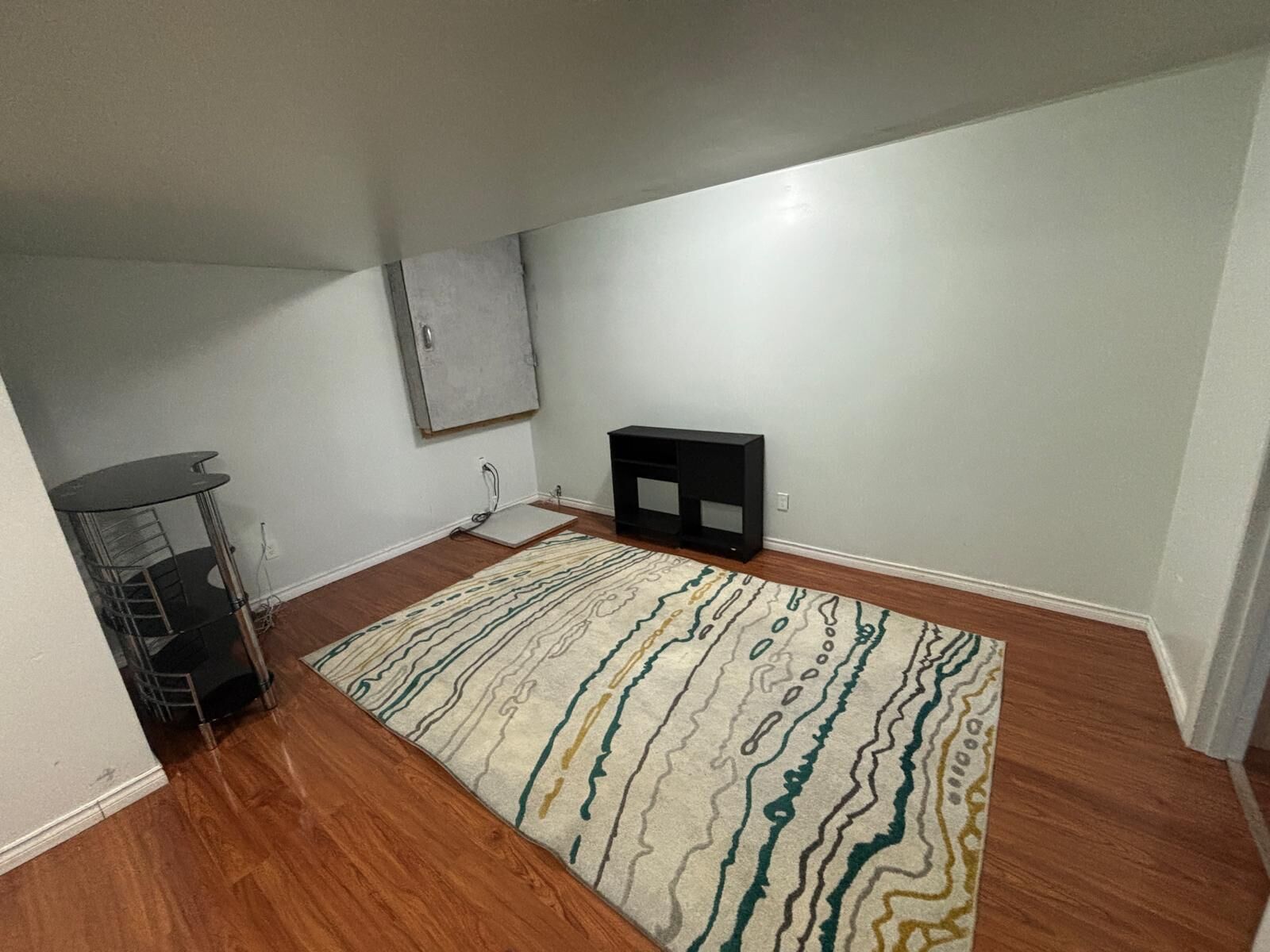 Une pièce avec un plancher en bois, un tapis à motifs, un meuble de rangement noir pour appareils électroniques et une étagère en métal.