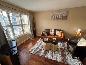 A living room with a brown leather sofa, two round coffee tables, a black armchair, and a large window with blinds. - Modern townhouse  offers the ideal blend of convenience and comfort, (Edmonton)