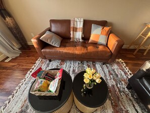 Un canapé en cuir marron agrémenté de coussins à motifs, une petite table noire sur laquelle est posé un vase de fleurs, et une boîte de collations sur un tapis à motifs.