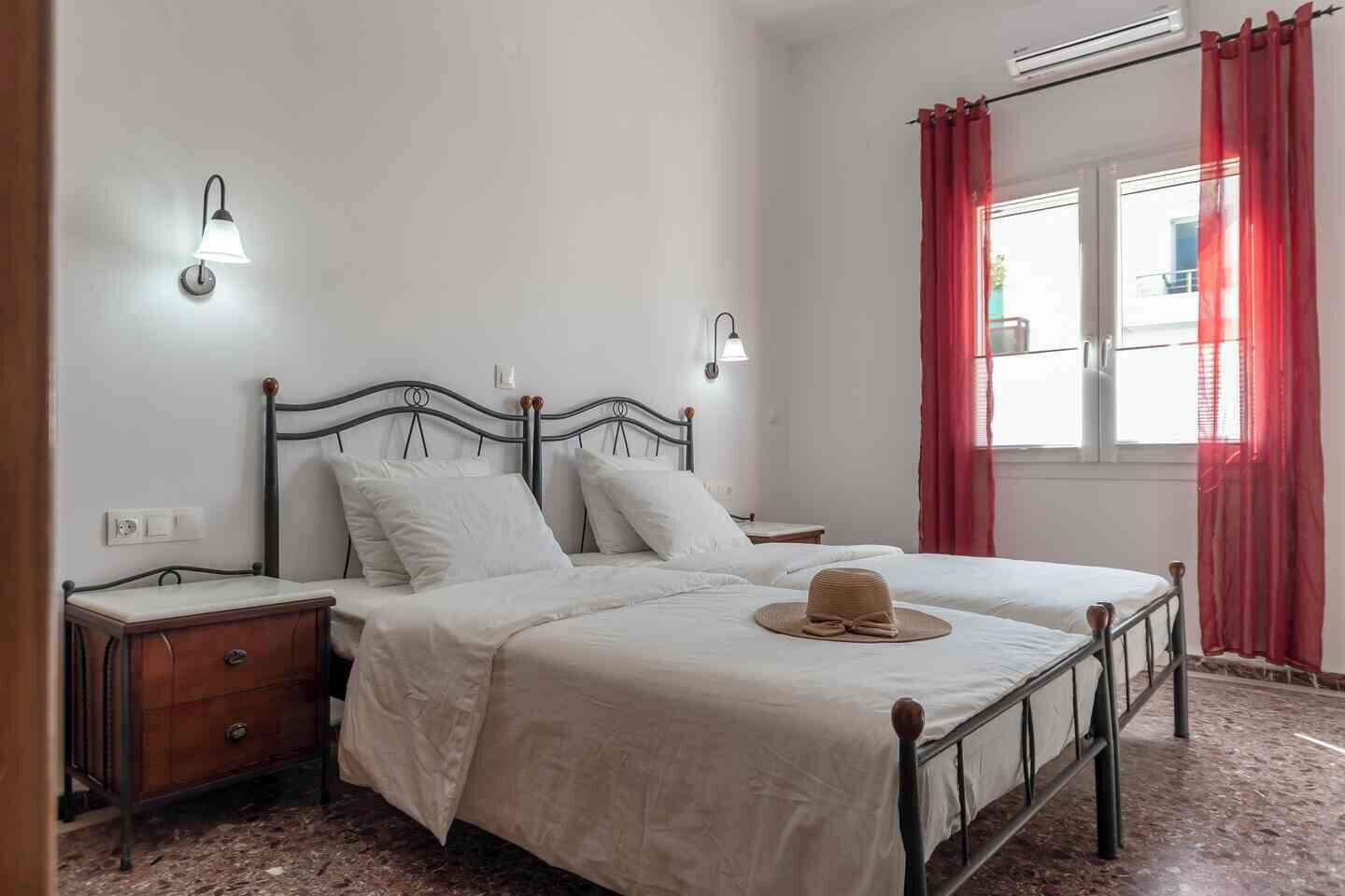A bedroom with a bed, two bedside tables, a wall-mounted lamp, a window with curtains, and a radiator.