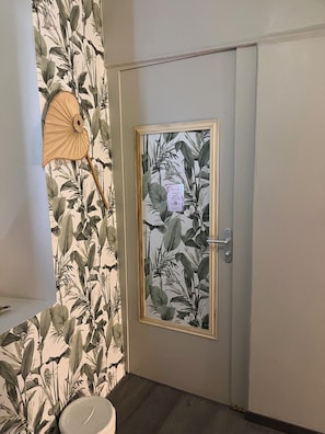 A room with a door featuring a botanical wallpaper, a wall-mounted fan, and a small shelf.