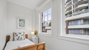 Una camera da letto con un letto di legno, un comodino, una lampada, una finestra vista su un edificio e un'opera d'arte incorniciata al muro.