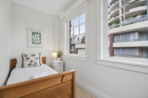 Ein Schlafzimmer mit einem hölzernen Bett, einem Nachttisch, einer Lampe, einem Fenster mit Blick auf ein Gebäude und einem gerahmten Kunstwerk an der Wand.