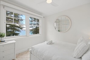 Ein Schlafzimmer mit einem Bett, einer Kommode, einem runden Spiegel und einem Fenster mit Blick auf Bäume und Wasser.