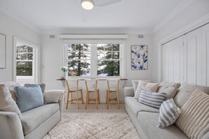 Ein gemütliches Wohnzimmer mit einem weißen Sofa, einem Couchtisch und einem Esstisch mit Stühlen. Ein großes Fenster bietet Blick auf Bäume und die Stadt.