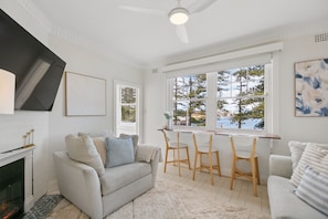 A living room with a fireplace, a sofa, a dining table with chairs, and a view of trees through large windows. - Manly Waterfront - Steps to Ferry, Water & Corso (Manly)