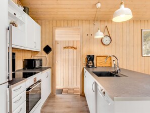 A modern kitchen with white cabinets, a stainless steel countertop, a built-in oven, and a dishwasher. There are two pendant lights, a clock, and a wooden door. - "Liina" - 3km from the sea by Interhome (Rømø)