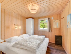 Une pièce aux murs en bois, un lit avec une literie blanche, une fenêtre avec des rideaux et une petite armoire.