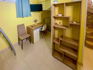 Ein Zimmer mit Schreibtisch, Stuhl, Fernseher und einem Holzregal.