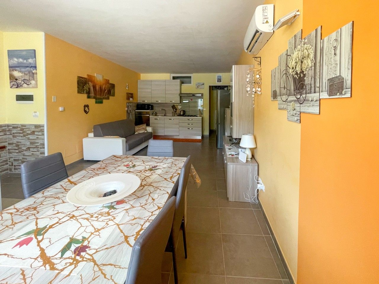 A dining area with a table set for two, a kitchen in the background, and a living area with a sofa and a TV.