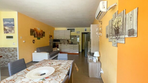 Un comedor con una mesa lista para dos personas, una cocina al fondo y una sala de estar con un sofá y un televisor.