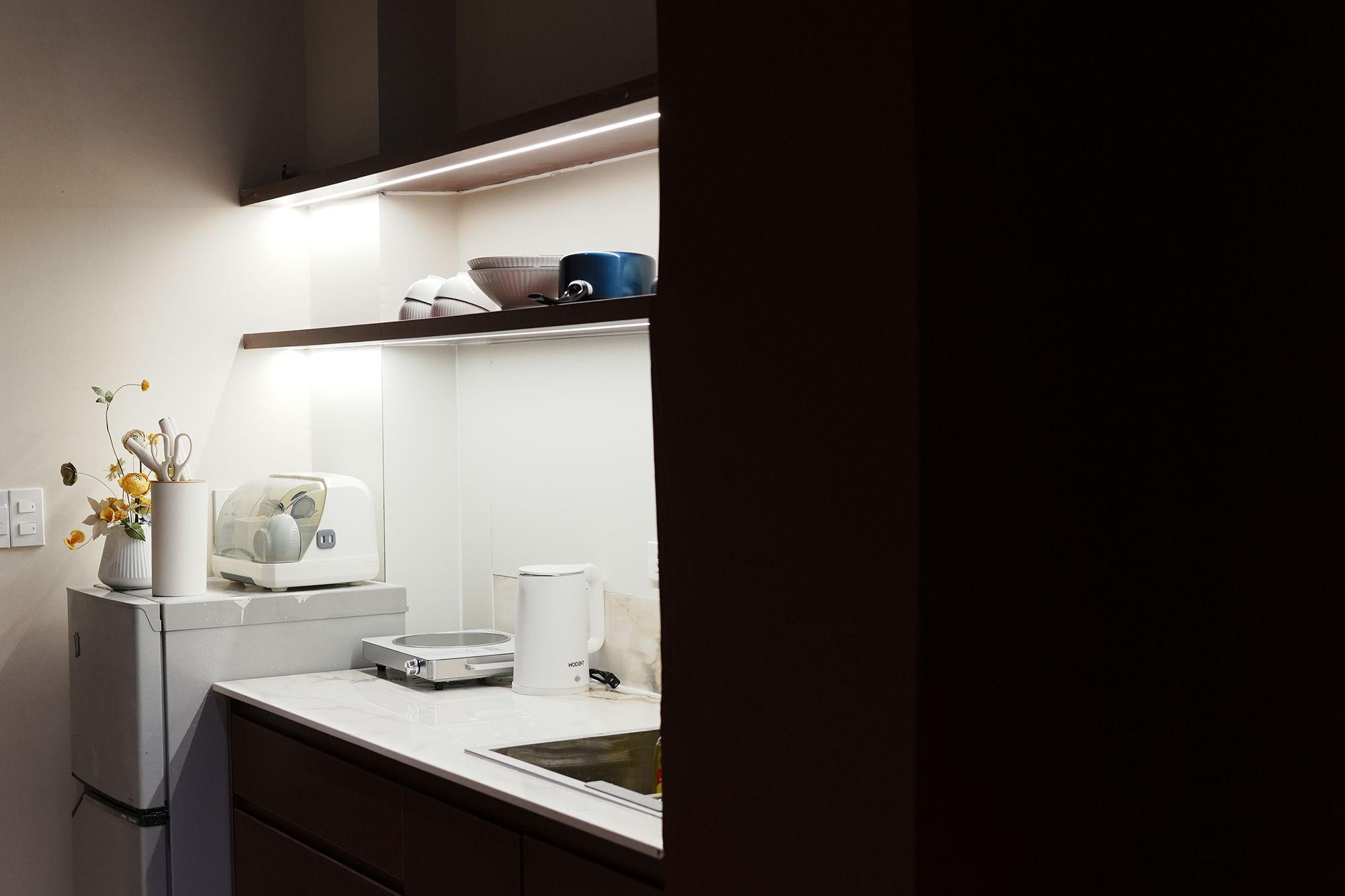 Een moderne keuken met een koelkast, magnetron en koffiezetapparaat op het aanrecht, en een plank erboven met keukengerei.