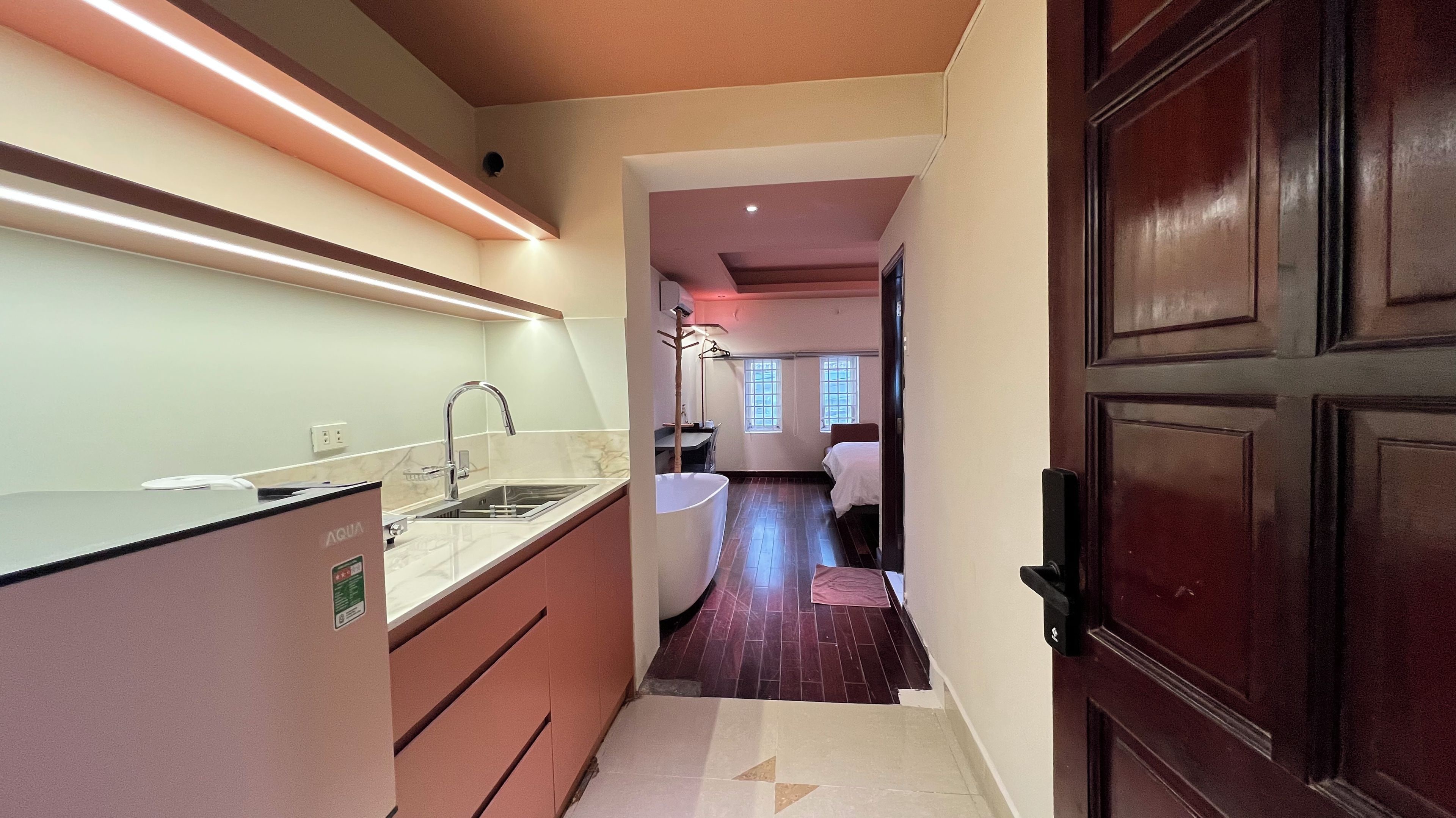 A narrow hallway leading to a bedroom with a wooden door, a kitchen area with a sink and countertop, and a window with blinds.