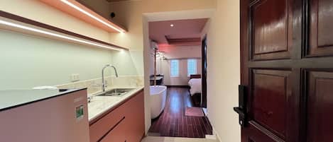 A narrow hallway leading to a bedroom with a wooden door, a kitchen area with a sink and countertop, and a window with blinds.