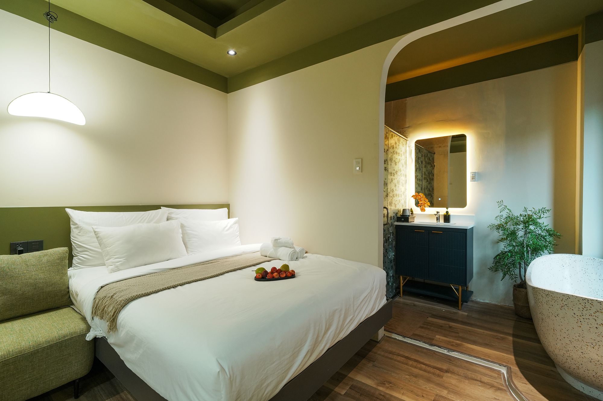 A hotel room with a bed, a sofa, a bathroom with a mirror, and a plant.