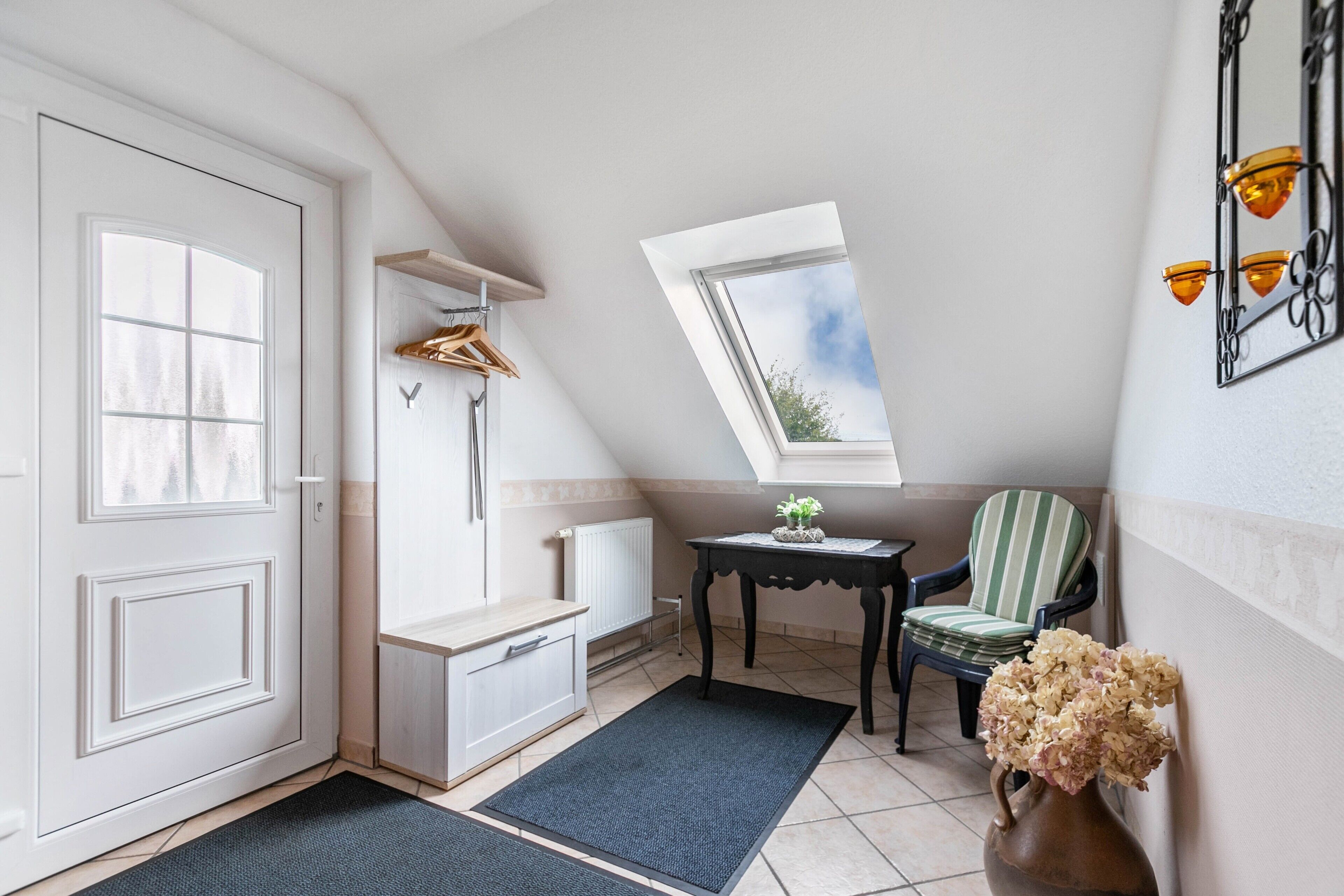 Um quarto com porta branca, um banco com cabide, um radiador, uma mesinha com uma planta, uma cadeira e um vaso com flores.