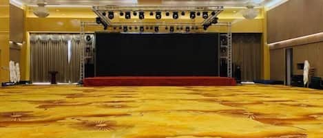 A large, carpeted hall with a stage, chandeliers, and a red carpet.