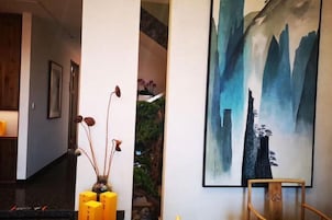 Un pasillo con una escalera, un cuadro de un paisaje montañoso y un jarrón con flores secas.