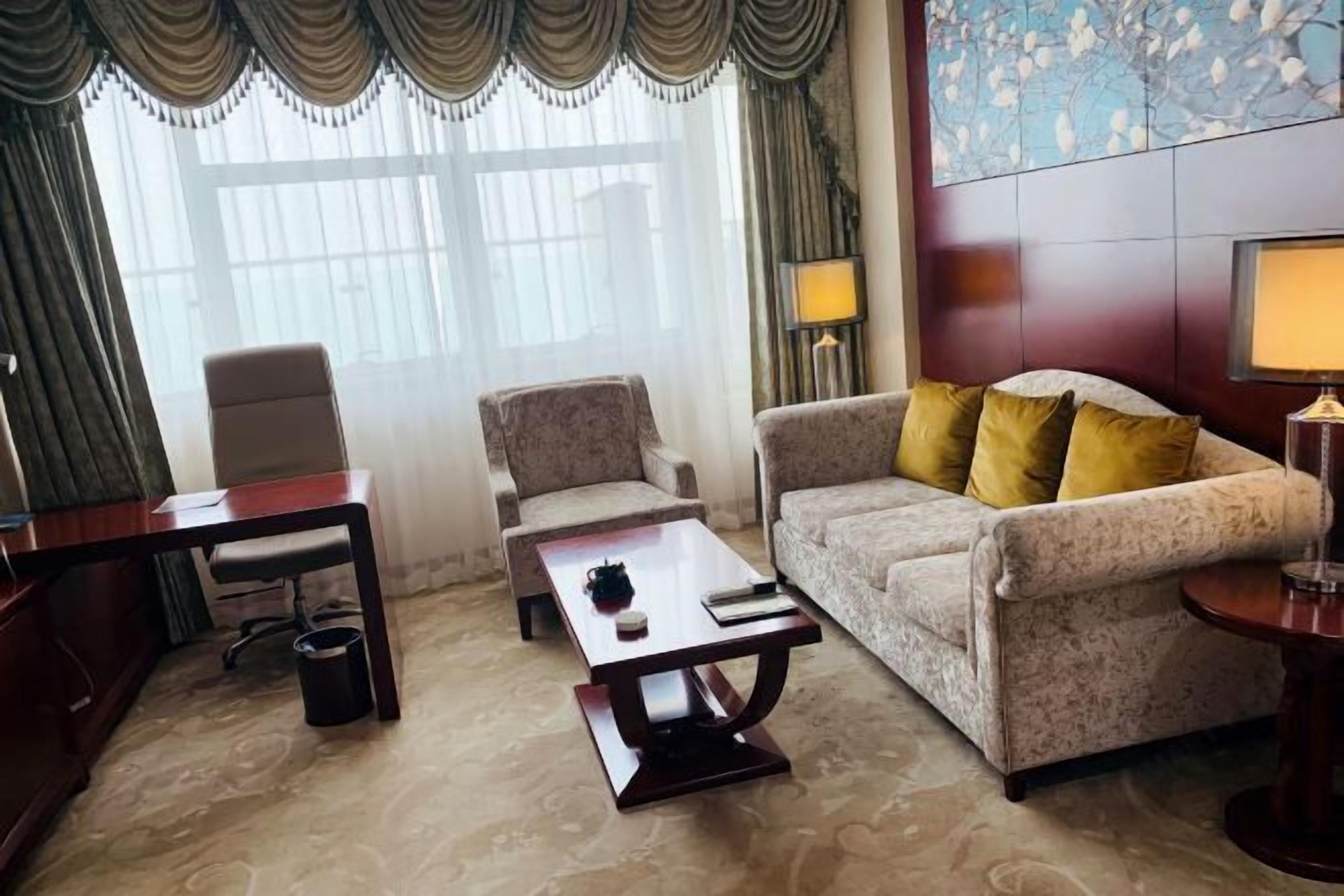 Quarto de hotel com sofá, poltrona, mesa de centro, escrivaninha e luminárias.