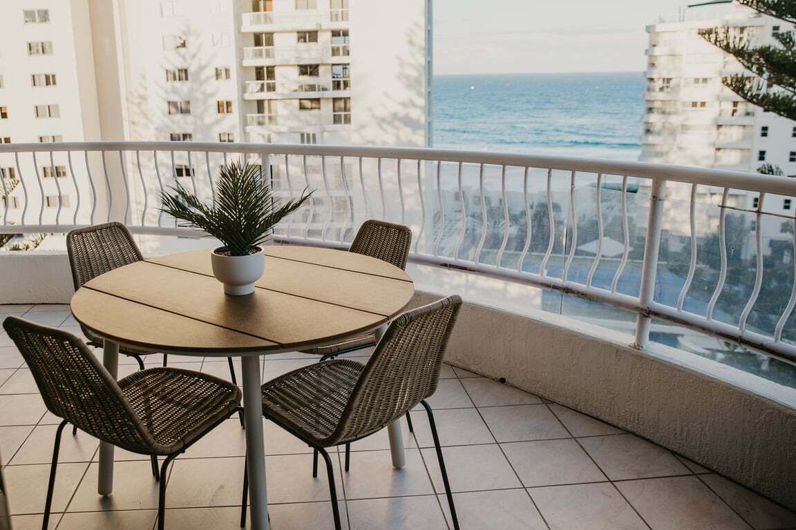 A round table with four chairs and a potted plant on a balcony with a view of the sea and buildings.