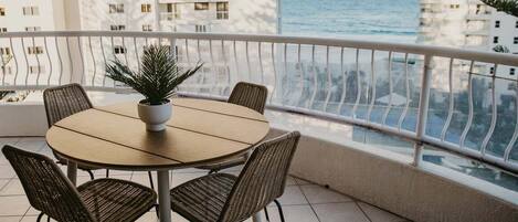 A round table with four chairs and a potted plant on a balcony with a view of the sea and buildings.