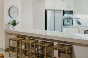 A modern kitchen with white cabinetry, stainless steel appliances, and a bar stool seating area. - Biarritz by the Sea | Resort Days & Ocean Views (Gold Coast)