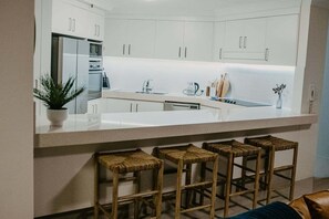 A modern kitchen with white cabinets, a central island with stools, and a built-in oven. - Biarritz by the Sea | Resort Days & Ocean Views (Gold Coast)